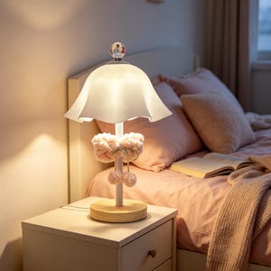 Può includere: Una lampada da tavolo con un paralume bianco a forma di campana svasata, un terminale in cristallo trasparente e uno stelo bianco. Lo stelo è decorato con un rivestimento in tessuto morbido, soffice e rosa chiaro con due piccoli pon-pon.