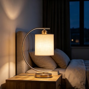May include: A table lamp with a curved brass-toned metal arm and a cylindrical beige fabric shade. The lamp emits a warm, soft glow and features a circular base, resting on a wooden surface next to a stack of books in a bedroom setting.