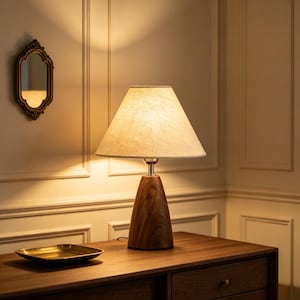 Könnte beinhalten: Eine Tischlampe mit einem konischen Holzfuß mit natürlicher Holzmaserung und einem beigen Stoffschirm. Die Lampe strahlt ein warmes Licht aus, das die Umgebung erhellt. Ein kleines, leeres, messingfarbenes rechteckiges Tablett steht daneben.