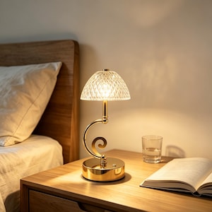 May include: A gold-toned table lamp featuring a circular base, a curved stem, and a translucent, dome-shaped shade with a diamond-cut pattern. The lamp emits a warm, soft glow, illuminating the surrounding area with a subtle, textured light effect.