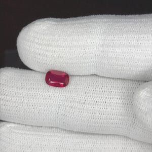 以下が含まれることがあります： クッションカットが施されたルビーの宝石。深く鮮やかな赤色をしており、光を反射するファセット面が特徴です。宝石は小さく、角が丸みを帯びた長方形で、高い透明度と全体にわたる鮮烈で濃い赤色の色調を示しています。