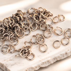 Könnte beinhalten: Eine Ansammlung von metallischen Biegeringen in Roségold-Optik. Die kleinen, kreisförmigen Schmuckverbinder sind verstreut, wobei einige zu einer kettenartigen Struktur verbunden sind und andere einzeln liegen, jeweils mit einer kleinen Öffnung.