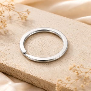 Puede incluir: Una anilla circular de metal plateado con un acabado pulido y reflectante. El anillo presenta un diseño de doble bucle en espiral, lo que permite enganchar llaves u otros objetos. La superficie del metal es lisa y de color metálico uniforme.