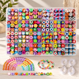 Könnte beinhalten: Eine durchsichtige Kunststoff-Sortierbox gefüllt mit bunten Tonperlen in Formen wie Sternen, Herzen, Tieren, Früchten und Smileys. Darunter liegen lose Perlen in einem Regenbogenbogen, zwei Perlenarmbänder und zwei Rollen mit durchsichtigem Gummiband.
