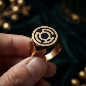 May include: A gold-toned signet ring featuring a circular face with a black inlay. The inlay displays a stylised, symmetrical emblem consisting of a central circle surrounded by four curved, interconnected segments forming a geometric pattern.