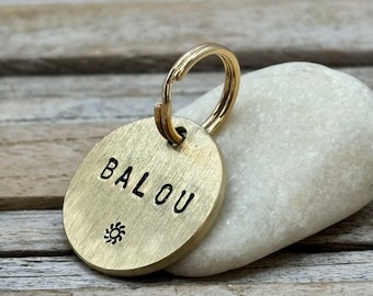 Op de afbeelding: Een ronde penning voor huisdieren van geborsteld messing met de naam BALOU in zwarte hoofdletters gestempeld, voorzien van een klein zon-icoontje onder de tekst en een goudkleurige metalen ring aan de bovenste lus.
