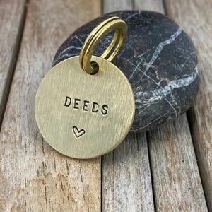 Könnte beinhalten: Ein kreisförmiger Messinganhänger mit gebürsteter Oberfläche und einem goldfarbenen Spaltring. Das Wort DEEDS ist in Großbuchstaben in die Mitte geprägt, mit einem kleinen Herzsymbol, das direkt unter dem Text auf der Vorderseite eingeprägt ist.