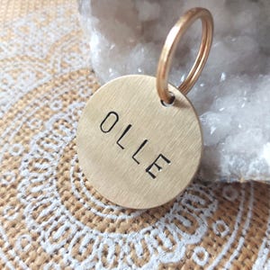 Op de afbeelding: Een ronde messing hanger met een geborstelde afwerking waarop de naam OLLE in zwarte hoofdletters is gestempeld. De hanger is bevestigd aan een bijpassende ronde messing sleutelring, wat een functioneel item vormt voor identificatie.
