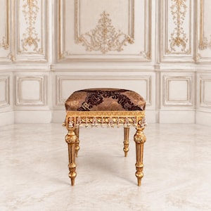 May include: A rectangular footstool featuring a dark brown and gold damask fabric seat with a gold fringe trim. The stool is supported by four ornate, carved gold-painted wooden legs with detailed fluting and bulbous accents at the top.