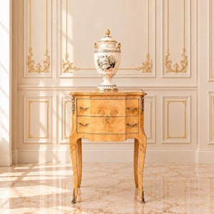 May include: A light-coloured wooden side table with two drawers, featuring ornate metal handles and curved legs, topped with a white porcelain urn decorated with gold accents and a painted scene of figures in period clothing.