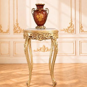 May include: A dark red glass vase with gold floral etchings and ornate handles sits atop a round table with a white marble top and a gold-finished, carved wooden base featuring curved legs and intricate floral relief details throughout.