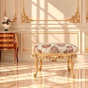 Op de afbeelding: Een ovale poef met een zitting van bruine en crèmekleurige damaststof, gebogen houten poten met een gouden afwerking en een decoratieve gouden franjerand versierd met kleine hangende kralen rond de onderkant van het gestoffeerde kussen.