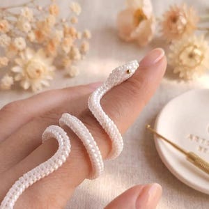 May include: A coiled snake ring crafted from small, uniform white beads. The snake features a single, tiny gold-colored bead for an eye. The body of the snake wraps around a finger in a spiral pattern, creating a flexible and textured appearance.