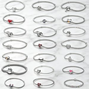 Puede incluir: Una colección de veintiuna pulseras de cadena de serpiente en tono plateado, cada una con un dije único. Los dijes incluyen varias formas como corazones, diseños inspirados en personajes, logotipos y símbolos, dispuestos en una cuadrícula.