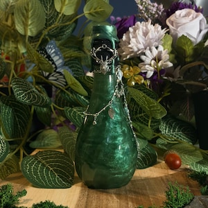 Peut inclure: Une bouteille en verre en forme de poire avec une finition vert émeraude scintillante. Le goulot est scellé avec de la cire foncée et orné d'une breloque en métal argenté représentant une tête de cerf, reliée à une fine chaîne argentée.