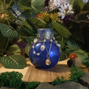 Peut inclure: Une fiole à potion bleue, ronde et brillante, dotée d'un col argenté. La fiole est ornée de fines chaînes dorées, de petites perles blanches artificielles et d'une breloque coquillage dorée contenant une perle blanche en son centre.