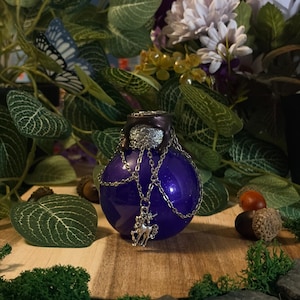 Può includere: Una bottiglia di vetro rotonda color viola intenso con finitura glitterata, dotata di un tappo marrone scuro e una catenella in metallo argentato drappeggiata attorno al collo. Un piccolo ciondolo a forma di unicorno pende dalla catena.