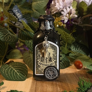 Può includere: Una bottiglia da speziale in vetro nero con collo glitterato argento e sigillo in ceralacca nera. L'etichetta presenta un'illustrazione di un cimitero e il testo APOTHECARY. Un ciondolo a forma di teschio d'uccello pende da una catena.