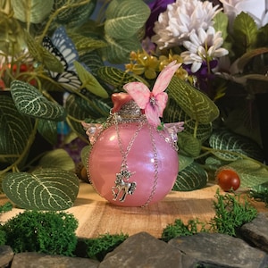 Può includere: Una bottiglia di vetro rosa perlato di forma rotonda con una catenella color argento avvolta attorno al collo. Un ciondolo a forma di unicorno argentato pende dalla catena. Un ornamento a farfalla rosa è fissato sulla parte superiore della bottiglia.