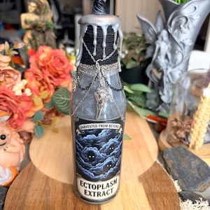 Può includere: Una bottiglia di vetro riempita di liquido grigio, sormontata da una statuetta argentata e cera argentata colante. Il collo è avvolto da spago nero e drappeggiato con catene argentate e un ciondolo a forma di teschio. L'etichetta riporta Harvested From Beyond Ectoplasm Extract.