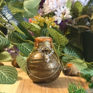 Peut inclure: Une bouteille sphérique de couleur bronze avec une finition métallique, dotée d'un bouchon scellé à la cire avec des coulures dorées. Une breloque tête de cerf argentée est fixée au col par des chaînes dorées et argentées délicates.