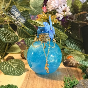 Peut inclure: Une bouteille en verre ronde remplie d'un liquide bleu scintillant, avec un bouchon scellé à la cire bleue. La bouteille est ornée de chaînes dorées, d'une petite clé en métal, d'une breloque hippocampe et d'une décoration papillon bleue.