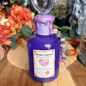 Può includere: Una bottiglia di pozione viola glitterata con un tappo trasparente a forma di cuore. Il collo è avvolto da un cordino viola con cera colante. L'etichetta riporta la scritta Potion d'Amour Elixir du Coeur Tendre con un cuore grafico.