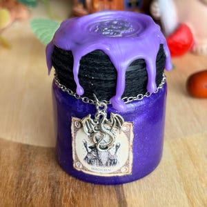 Peut inclure: Un pot en verre violet à la finition scintillante, surmonté de coulures de cire violette sur un couvercle enveloppé de corde noire. Une chaîne argentée entoure le col avec un pendentif dragon. Une étiquette rectangulaire porte le texte MAGICIEN.