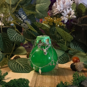 Può includere: Una fiala rotonda di colore verde brillante con un tappo di sughero sigillato con cera verde chiaro. La bottiglia è decorata con una catenina argentata con un ciondolo a forma di testa di cervo e piccoli ciondoli a foglia sul corpo in vetro.