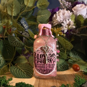 Può includere: Una bottiglia di vetro riempita con liquido rosa scintillante, sormontata da gocce di cera rosa. L'etichetta riporta LOVE POTION FOR AFFLICTIONS OF THE HEART AND ENSNARING OF THE SOUL MIX 1/2 WITH ROSEWATER. Una chiave argentata pende dal collo.