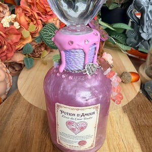 Puede incluir: Una botella de vidrio llena de líquido morado brillante, rematada con un tapón transparente en forma de corazón. El cuello está envuelto en hilo lavanda con cera rosa efecto goteo y pedrería. La etiqueta muestra el texto Potion d'Amour.