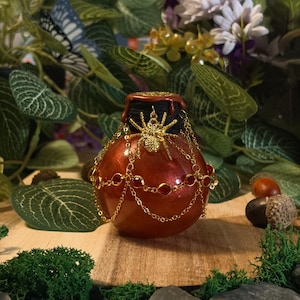 Può includere: Una boccetta per pozioni rotonda di colore arancione metallizzato con un ciondolo a forma di ragno dorato fissato al collo. La boccetta è decorata con delicate catenelle dorate e piccole perline rosse rotonde drappeggiate sul davanti, con un sigillo in cera scuro.