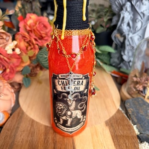 Peut inclure: Une bouteille en verre transparent remplie de liquide orange, avec une étiquette indiquant Monster Extract Chimera Fat Oil. Le goulot est entouré de corde noire avec de la cire jaune qui coule, orné d'une chaîne dorée et de cristaux rouges.