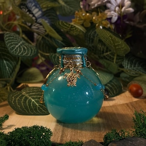 Può includere: Una bottiglia di vetro rotonda color verde acqua riempita con un liquido scintillante. Il collo è avvolto da un cordino argentato e sigillato con cera verde acqua. Una catenina dorata pende dal collo con un ciondolo a tartaruga e perline blu.