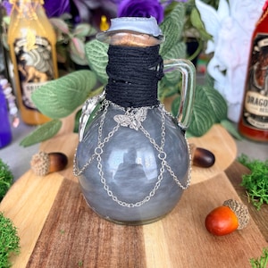 Può includere: Una bottiglia in vetro trasparente con manico, riempita con un liquido grigio vorticoso. Il collo è avvolto da uno spago nero e sigillato con un tappo di sughero. Una catena in metallo argentato avvolge il corpo con un ciondolo a forma di falena.