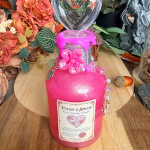 Puede incluir: Una botella de vidrio llena de líquido rosa brillante, rematada con un tapón transparente en forma de corazón. El cuello presenta gotas de cera rosa, una flor rosa y una etiqueta que dice Potion d'Amour Elixir du Coeur Tendre.