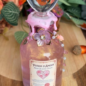 Può includere: Una bottiglia di vetro riempita con liquido rosa e oro scintillante, sormontata da un tappo trasparente a forma di cuore e gocce di cera rosa. L'etichetta riporta Potion d'Amour Elixir du Cœur Tendre, decorata con un ciondolo floreale e perline.
