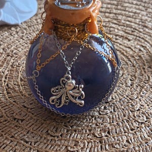 Peut inclure: Une bouteille en verre ronde remplie d'un liquide violet foncé, scellée avec de la cire orange portant un emblème argenté. La bouteille est ornée de fines chaînes dorées et argentées, et une breloque pieuvre argentée pend au centre du goulot.