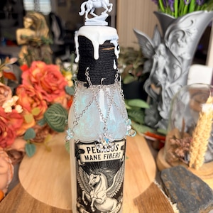 Può includere: Una bottiglia di vetro trasparente riempita di liquido iridescente, con un'etichetta che recita PEGASUS MANE FIBERS USED IN FLIGHT SPELLS. Il collo è avvolto da spago nero con gocce di cera bianca e una statuetta di Pegaso bianco, con catene argentate.