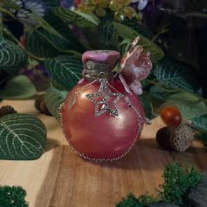 Peut inclure: Une bouteille en verre rose nacré de forme ronde, ornée d'une breloque étoile en argent représentant une petite fée. Le goulot est entouré de fil doré, scellé par de la cire rose et décoré d'une fleur en papier pailletée à plusieurs pétales.