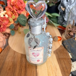 Peut inclure: Une bouteille en verre transparent remplie d'un liquide bleu scintillant, surmontée d'un bouchon en forme de cœur et d'un sceau en cire grise. Elle porte une étiquette Potion d'Amour Elixir du Coeur Tendre, une breloque aile et des perles.