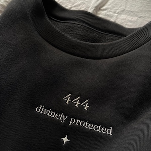 Könnte beinhalten: Ein schwarzes Sweatshirt mit Rundhalsausschnitt und weißer Stickerei mit der Aufschrift 444 divinely protected, platziert über einem kleinen vierzackigen Sternsymbol. Das Kleidungsstück hat einen gerippten Kragen und eine weiche Fleece-Innenseite.