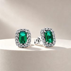 May include: A pair of silver stud earrings featuring a rectangular emerald-cut green gemstone at the center, surrounded by a halo of small, round, clear white stones. The earrings have a polished metallic finish and a secure post-back mechanism.