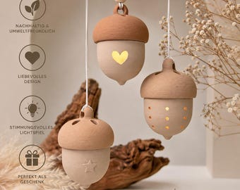 Könnte beinhalten: Drei hängende eichelförmige Ornamente aus matter, hellbrauner Keramik. Jedes Ornament hat eine dunkelbraune Kappe und ausgestanzte Formen wie ein Herz, einen Stern und kleine Punkte, durch die das Licht einer internen Quelle scheint.