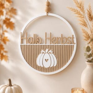 Könnte beinhalten: Ein kreisförmiger Wandbehang mit weißem Rahmen und beigefarbenem Lamellenhintergrund. Der Schriftzug "Hallo Herbst" steht in beigen Buchstaben über einem weißen Kürbis-Symbol mit einem kleinen herzförmigen Ausschnitt in der Mitte. Mit Holzperle und Schnur.