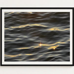 Puede incluir: Una impresión de arte mural enmarcada que muestra una masa de agua oscura y ondulada con la luz dorada del sol reflejándose en las crestas de las olas. La imagen está encerrada en un marco negro fino y sencillo, creando un contraste marcado.