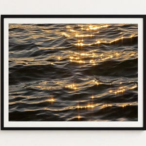Può includere: Una stampa artistica da parete incorniciata che raffigura uno specchio d'acqua scuro e increspato al tramonto. Le onde nero profondo e blu marino sono illuminate dalla luce dorata del sole, creando riflessi luminosi sulla superficie.