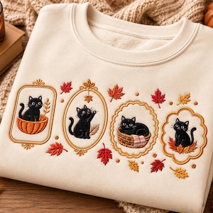 Può includere: Una felpa girocollo color crema con quattro cornici ovali ricamate, ognuna contenente un gatto nero. I gatti sono raffigurati seduti in una zucca, mentre cercano di afferrare una foglia, riposano in un cesto e siedono su foglie autunnali.