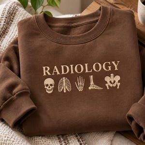 Puede incluir: Una sudadera marrón de cuello redondo con la palabra RADIOLOGY bordada en hilo color crema. Debajo del texto hay cinco pequeñas y detalladas ilustraciones bordadas en color crema de partes del esqueleto humano: un cráneo, caja torácica, mano, pie y pelvis.