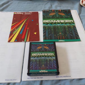 Könnte beinhalten: Ein klassisches Beamrider-Videospielmodul für das Atari 2600, zusammen mit der originalen Bedienungsanleitung und einem Activision-Videospielkatalog. Das Modul und das Handbuch zeigen futuristische grüne und blaue geometrische Grafiken.